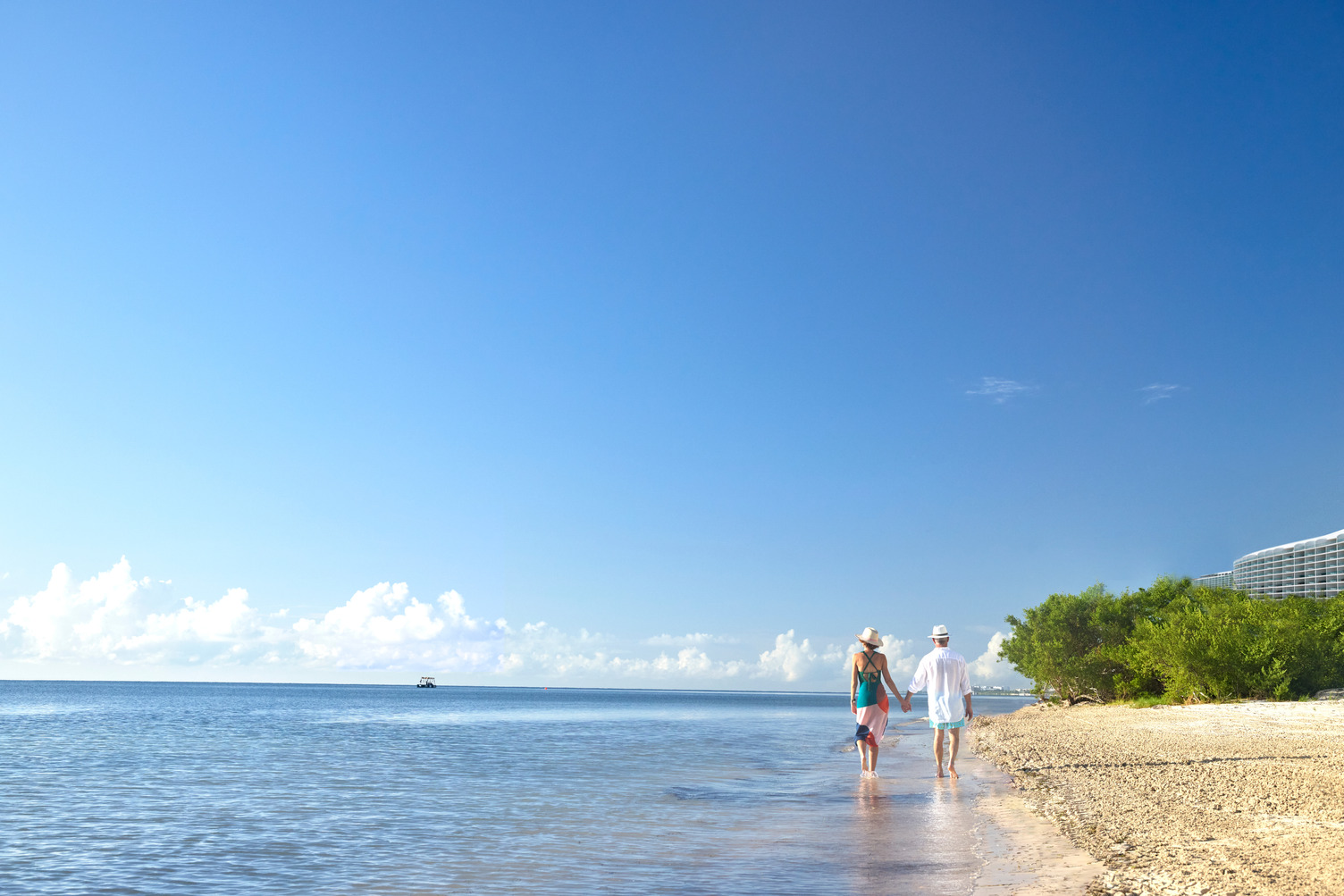 Couple walking along a sandy beach beside calm ocean waters under a clear blue sky.