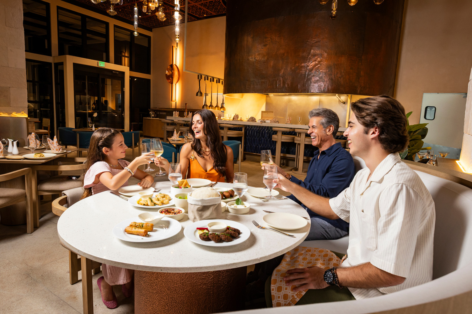Family dining in a modern restaurant, smiling and toasting with water glasses over a table of assorted dishes.