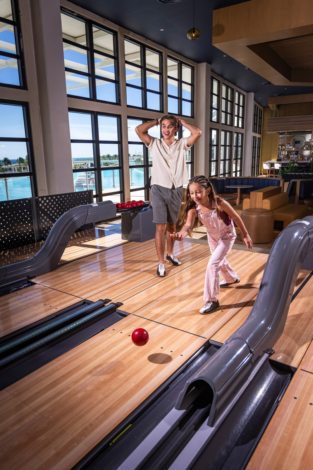 Man and girl enjoy bowling in a bright alley with large windows overlooking a pool area.