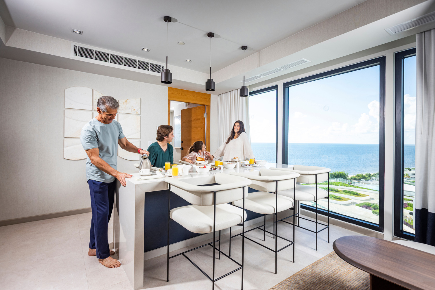 Family enjoying breakfast in a bright, modern resort room with ocean view through floor-to-ceiling windows.