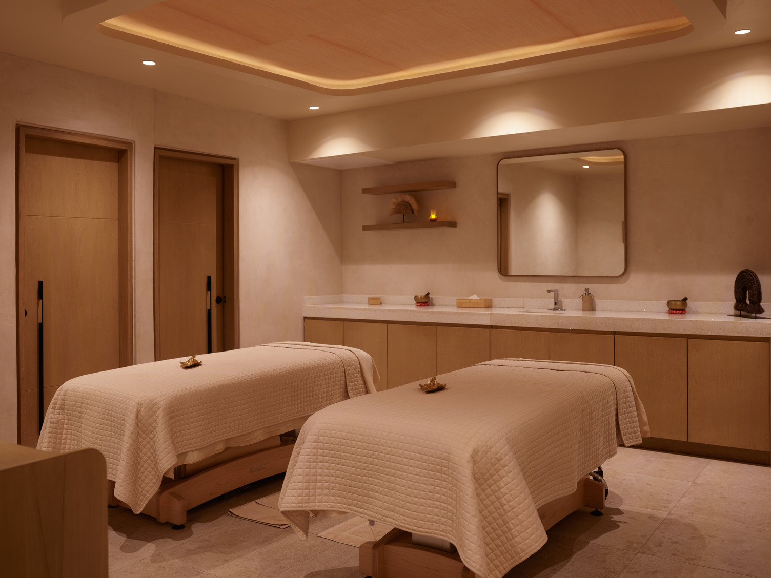 Serene spa room with two massage tables, soft lighting, white linens, and a large mirror on the back wall.