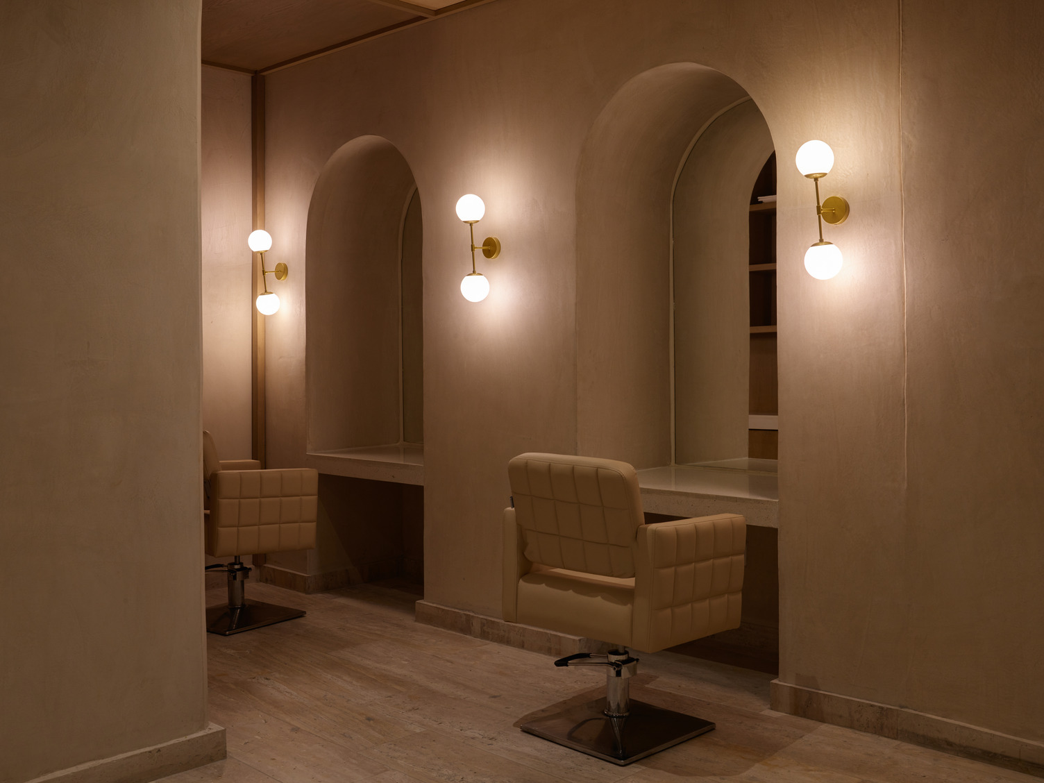 Minimalist spa interior with beige walls, arched mirrors, and cushioned salon chairs illuminated by globe wall lights.