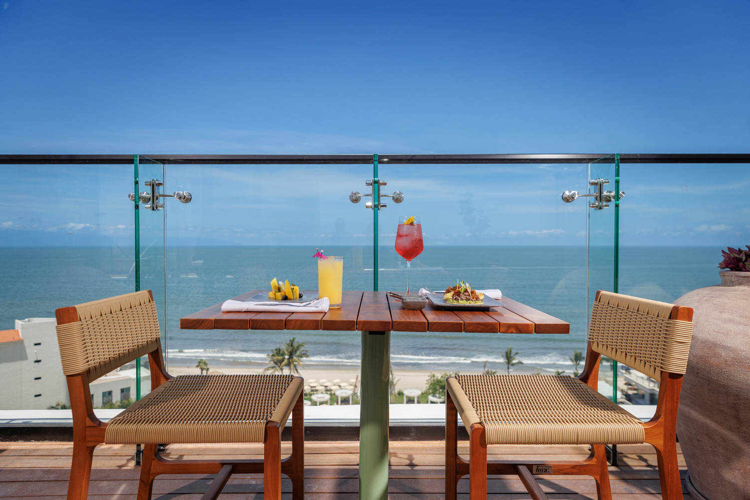 Outdoor restaurant table set for two with tropical drinks, overlooking the ocean under a clear blue sky.