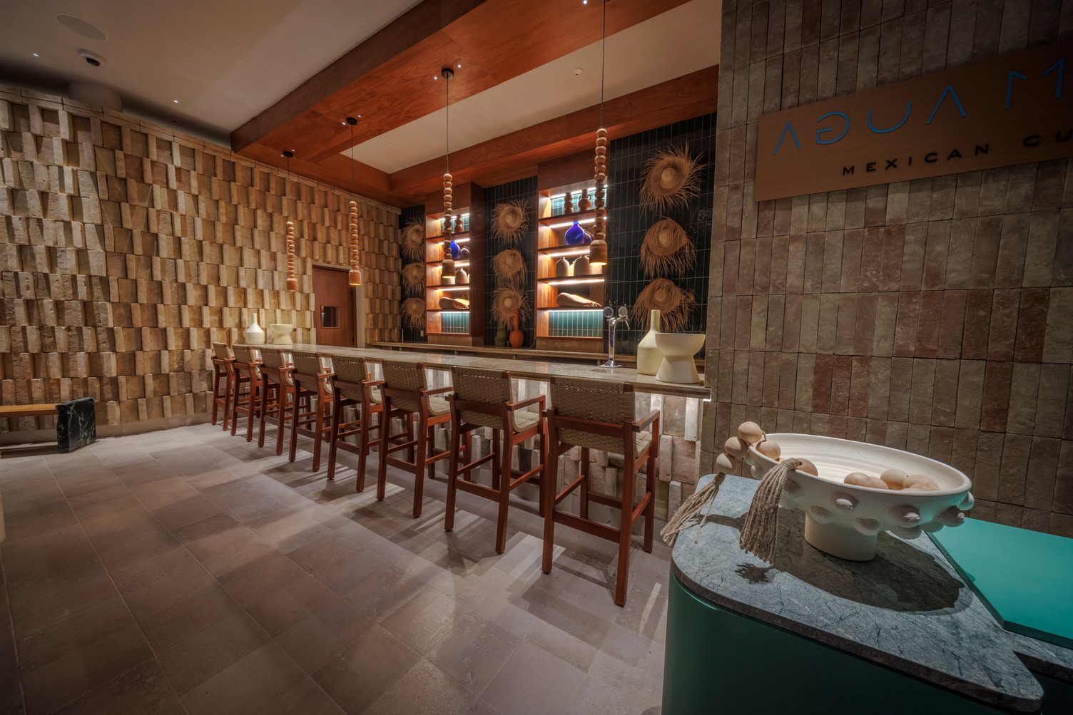 Elegant restaurant interior with a long bar, wooden chairs, patterned walls, and decorative ceiling lights.