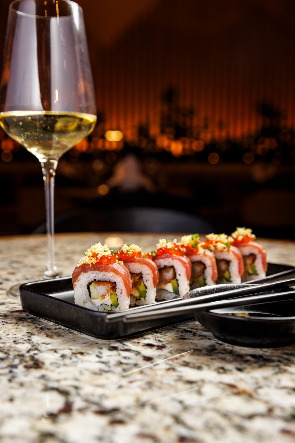 Sushi rolls with garnish on a black plate, beside a glass of white wine on a speckled counter.