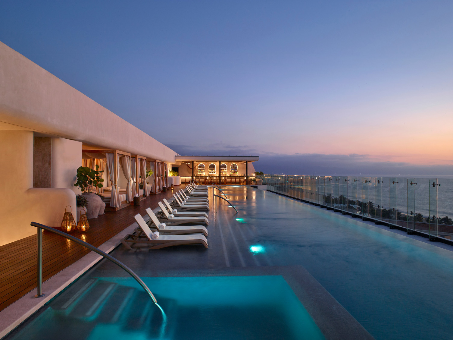 Rooftop infinity pool at sunset with lounge chairs and ocean views, lined by modern cabanas and glass railing.