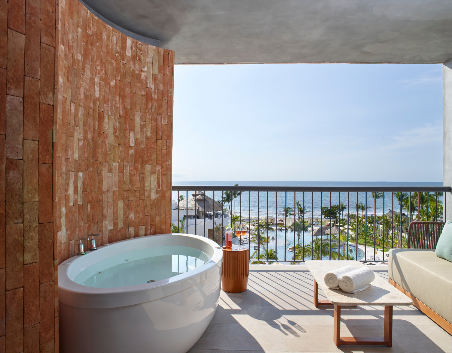 Balcony with ocean view, featuring a round tub, wicker table, and pool area surrounded by palm trees.