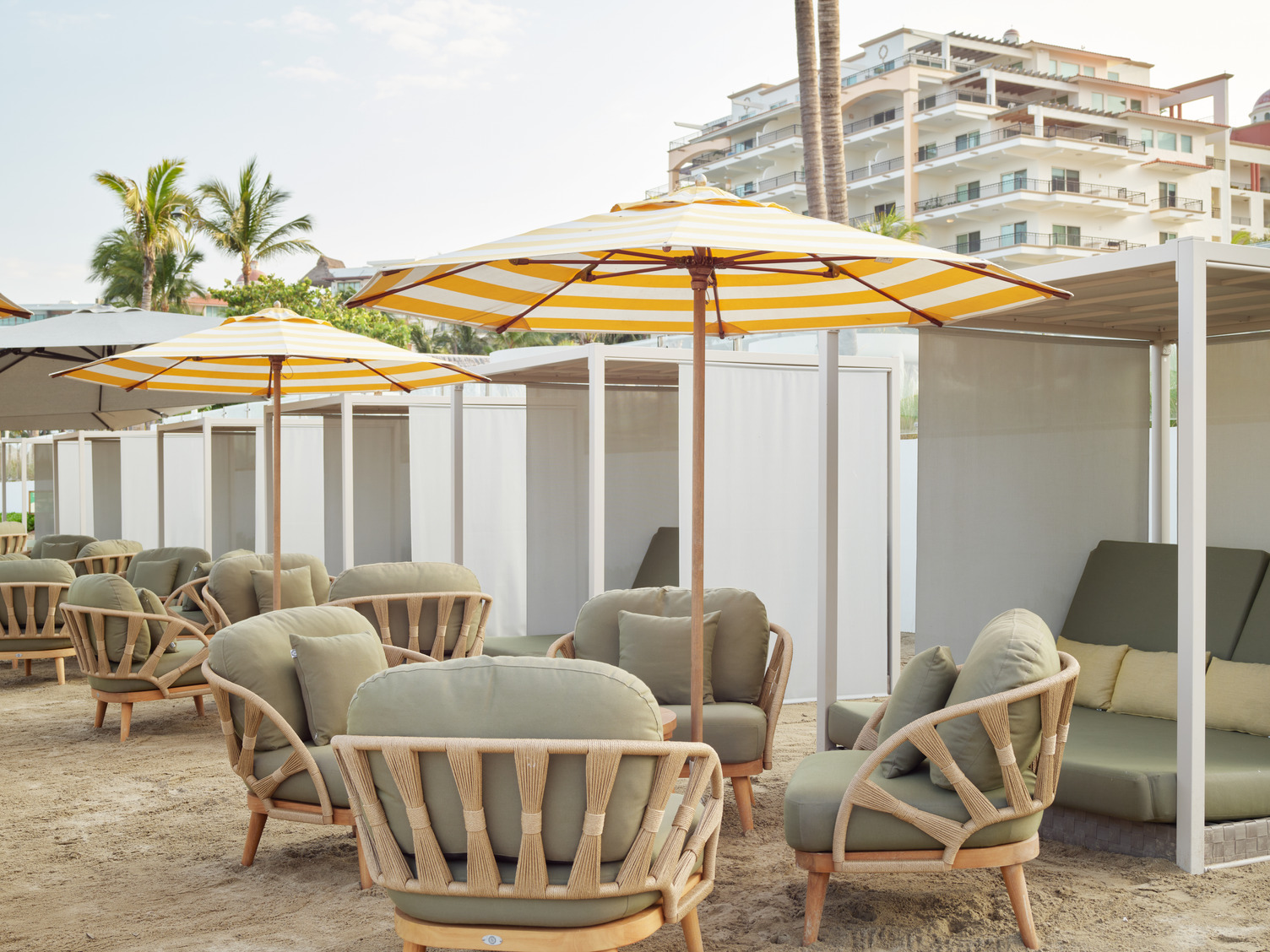 Outdoor lounge area with wicker chairs, yellow-striped umbrellas, and cabanas set on a sandy beach near a building.