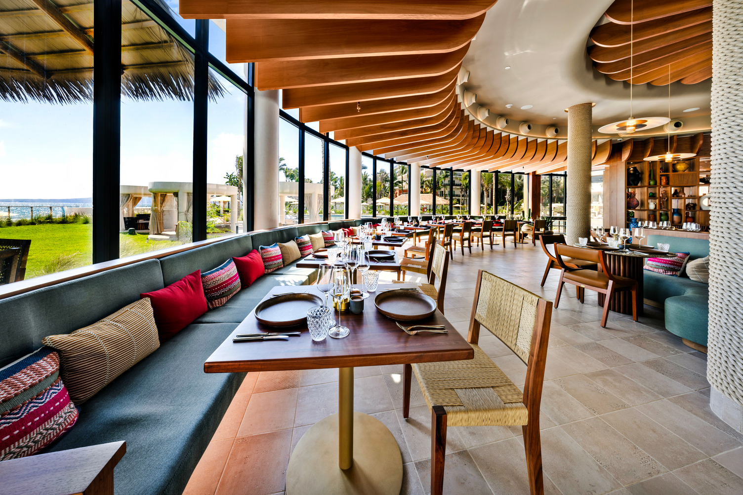 Elegant restaurant interior with ocean view, featuring wooden ceiling, colorful seating, set tables, and large windows.