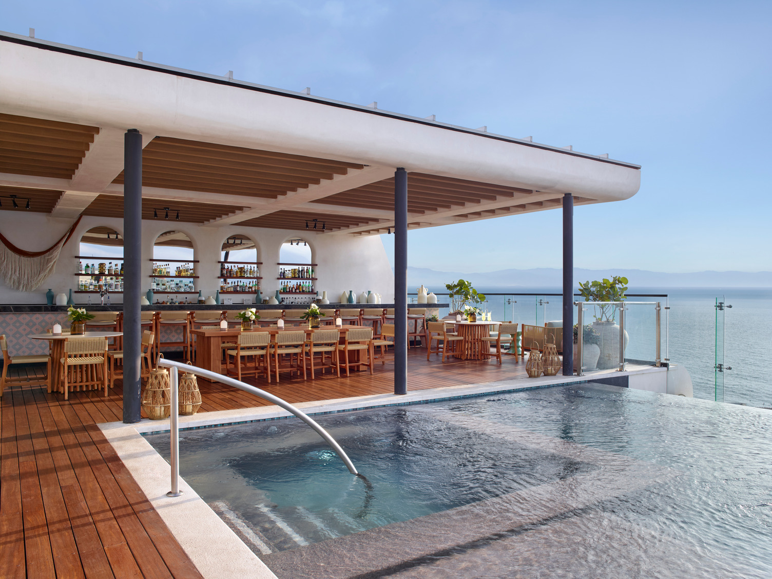 Open-air bar with wooden tables and chairs, infinity pool, glass railing, ocean and mountain views at Alto 20° 105° bar.