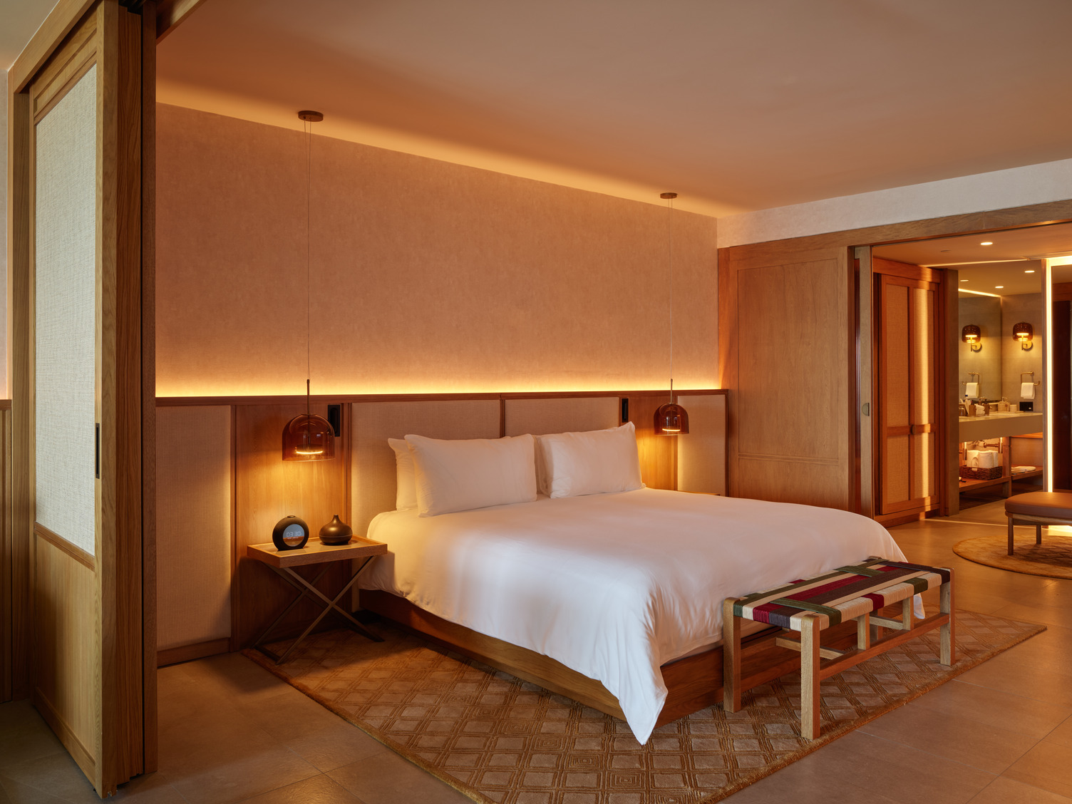 Luxury hotel suite with a king-sized bed, warm lighting, wooden decor, and a view into the modern bathroom area.