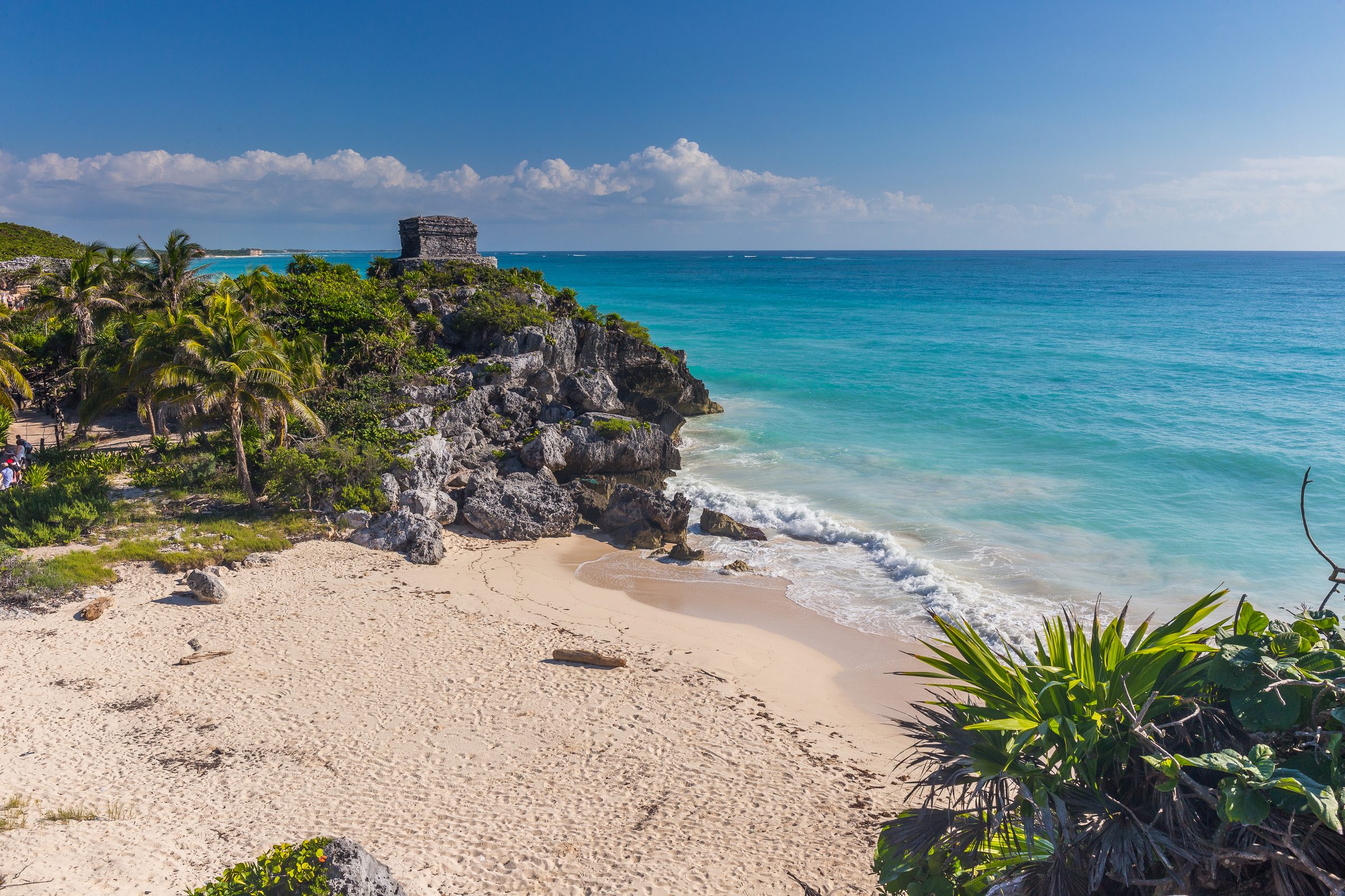 Playa de Tulum con agua turquesa, arena y ruinas antiguas en la cima de un acantilado rocoso bajo un cielo azul claro.