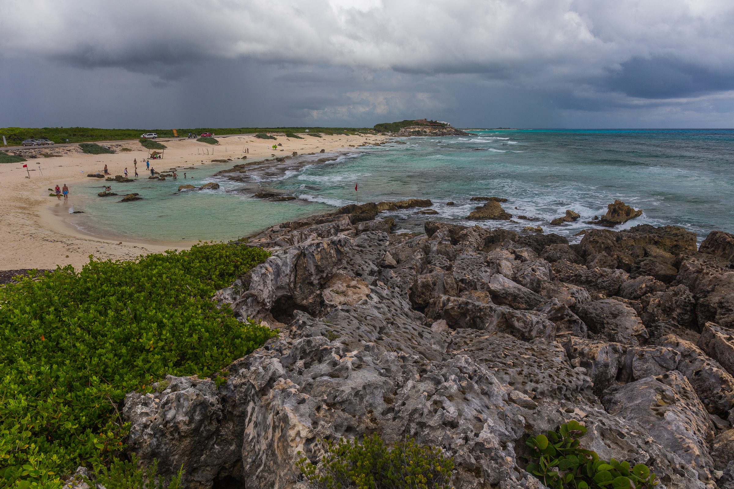 Costa rocosa y playa arenosa en Isla Mujeres, México, con vegetación, personas y cielos nublados.