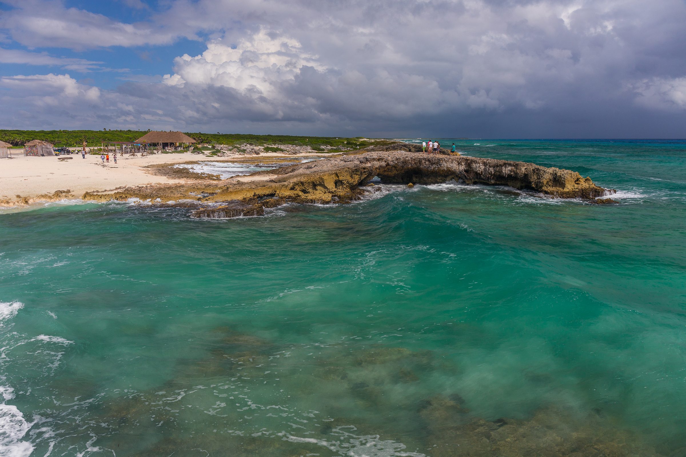 Rocky formation extends into turquoise Caribbean Sea under a cloudy sky, with huts and people on a sandy beach.