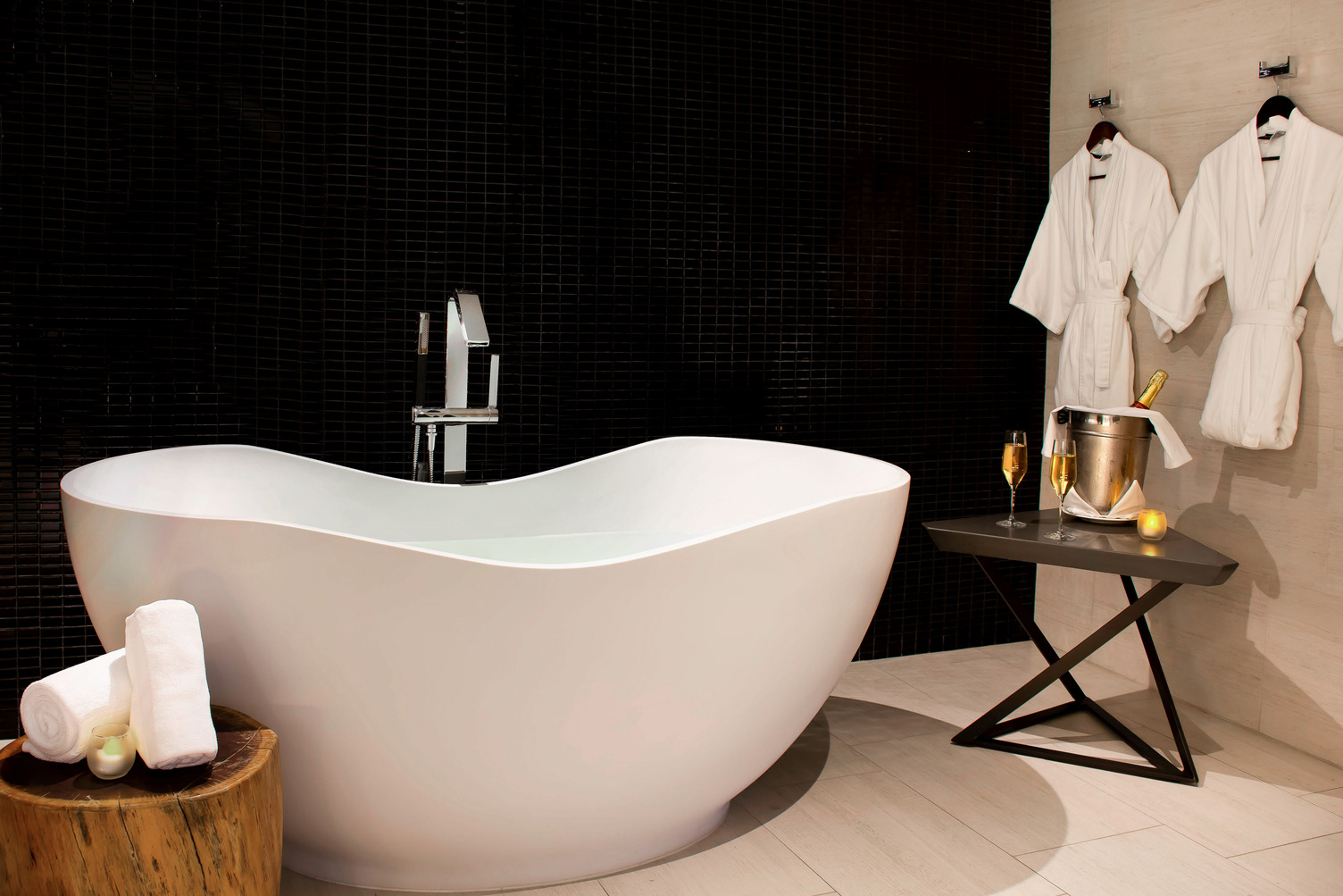 Lujosa tina de baño blanca con un fondo de azulejos oscuros, dos batas, champán y toallas en un banco de madera.