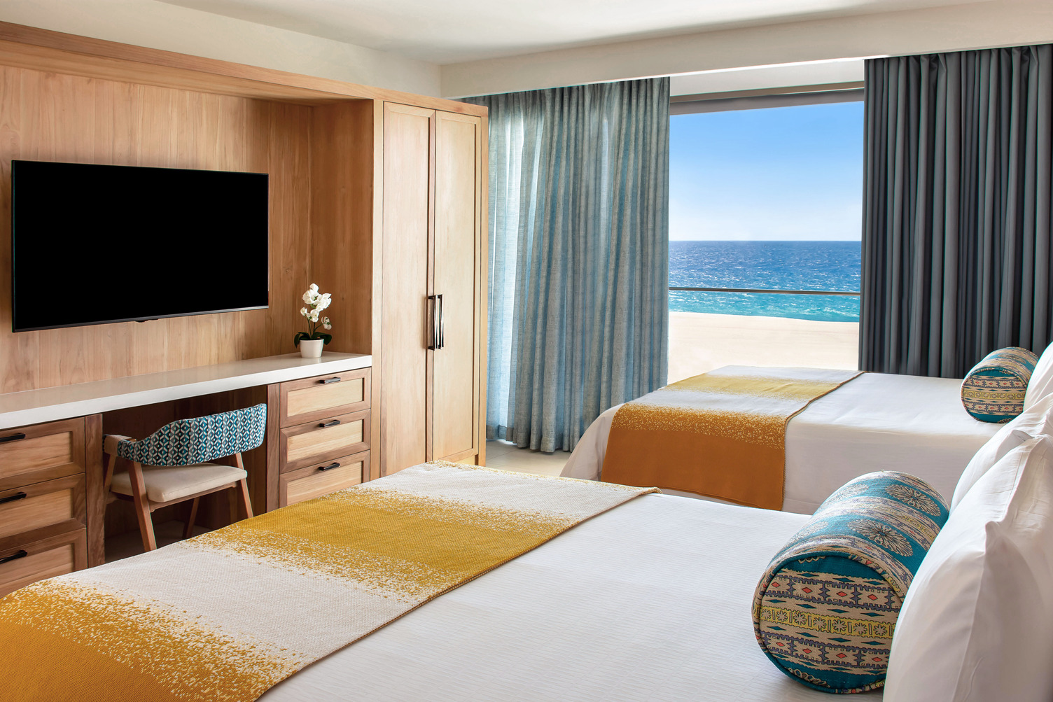 Oceanfront bedroom with two double beds, vibrant pillows, a wooden desk, TV, and large window view of the sea.