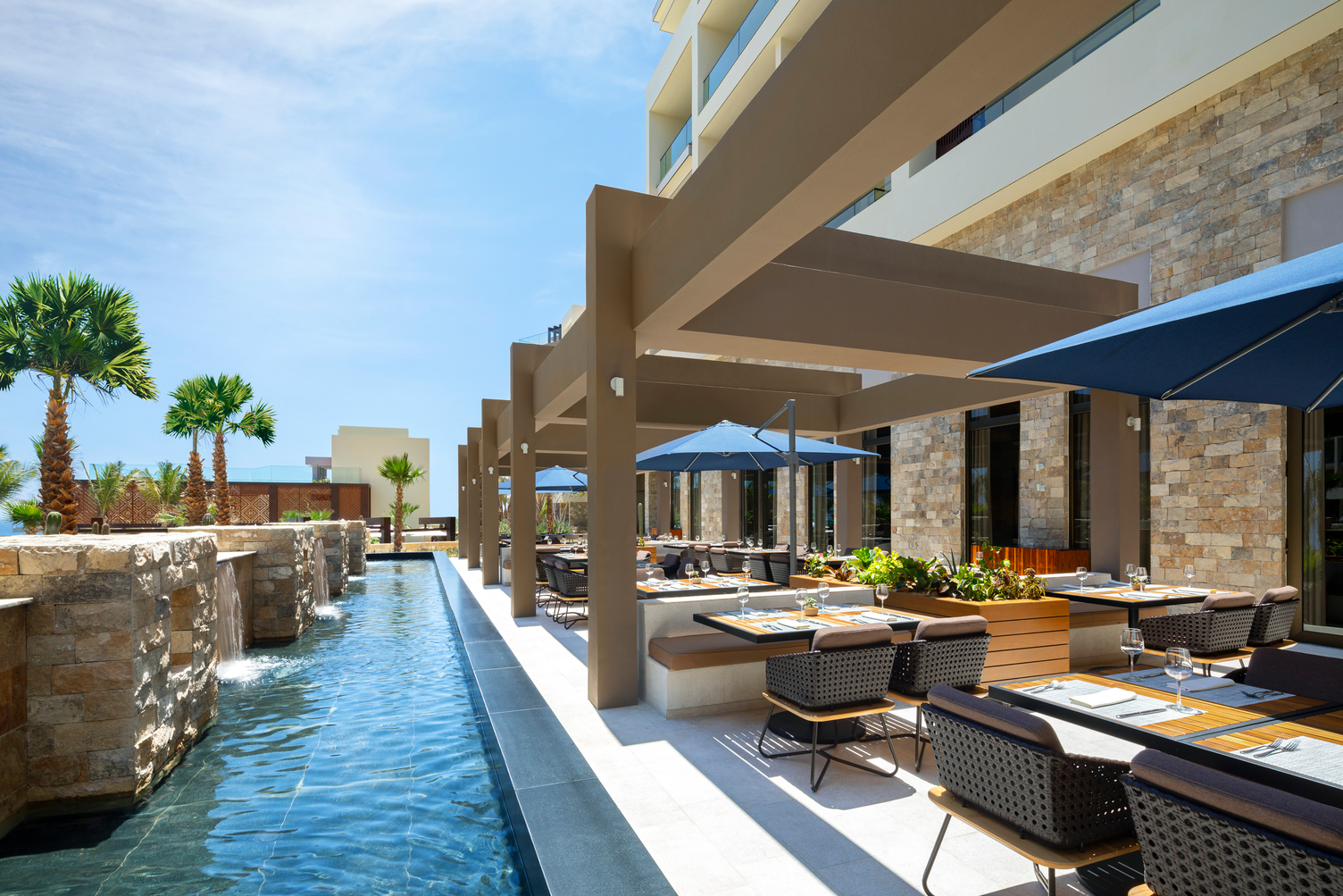 Terrace dining area with shaded tables, a pergola, and a water feature at a resort, HRHLC_CIAO restaurant.