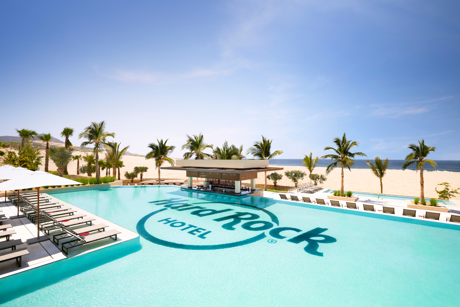 Sunny poolside with Hard Rock Hotel logo, surrounded by palm trees; beach and blue sky in the background.