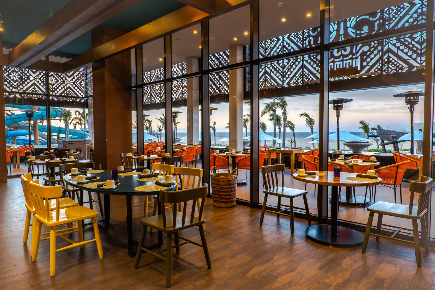 Restaurant interior with wooden tables and chairs, large windows overlooking a scenic ocean view at sunset.