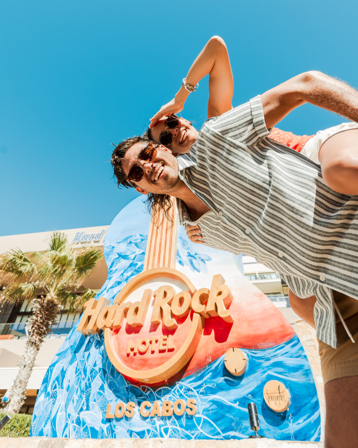 Pareja posa de manera juguetona frente al letrero del Hard Rock Hotel Los Cabos con diseño de guitarra bajo un cielo azul cla