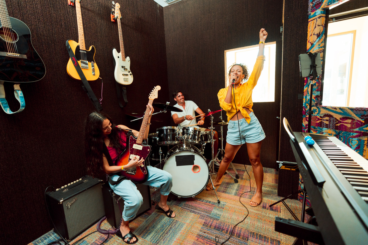 Tres personas en una sala de música con guitarras en la pared; una toca la guitarra, otra la batería y una canta con entusias