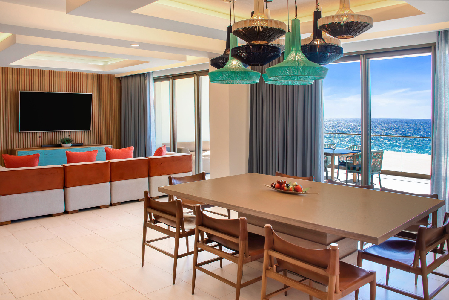 Área de estar moderna con vista al océano, con mesa de comedor de madera, luces colgantes coloridas, sofá y televisión de pan