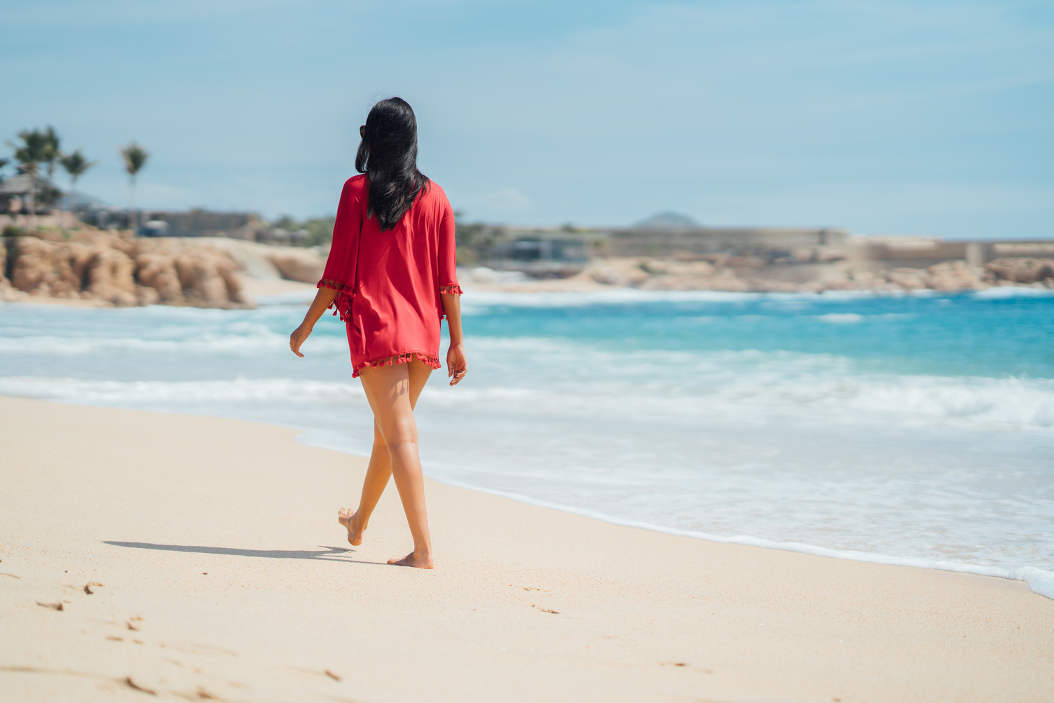 Mujer con un pareo rojo caminando por una playa soleada y arenosa con olas suaves en Playa Chileno, Los Cabos.