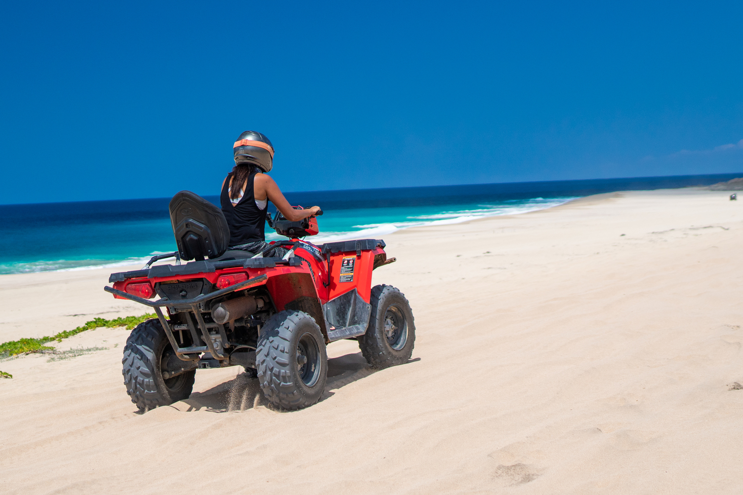Persona conduciendo un ATV en una playa de arena con el océano azul brillante y cielo despejado en Los Cabos.