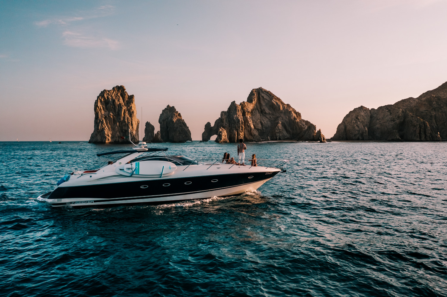 Yate de lujo navegando cerca de formaciones rocosas al atardecer en Los Cabos, con un océano tranquilo de fondo.