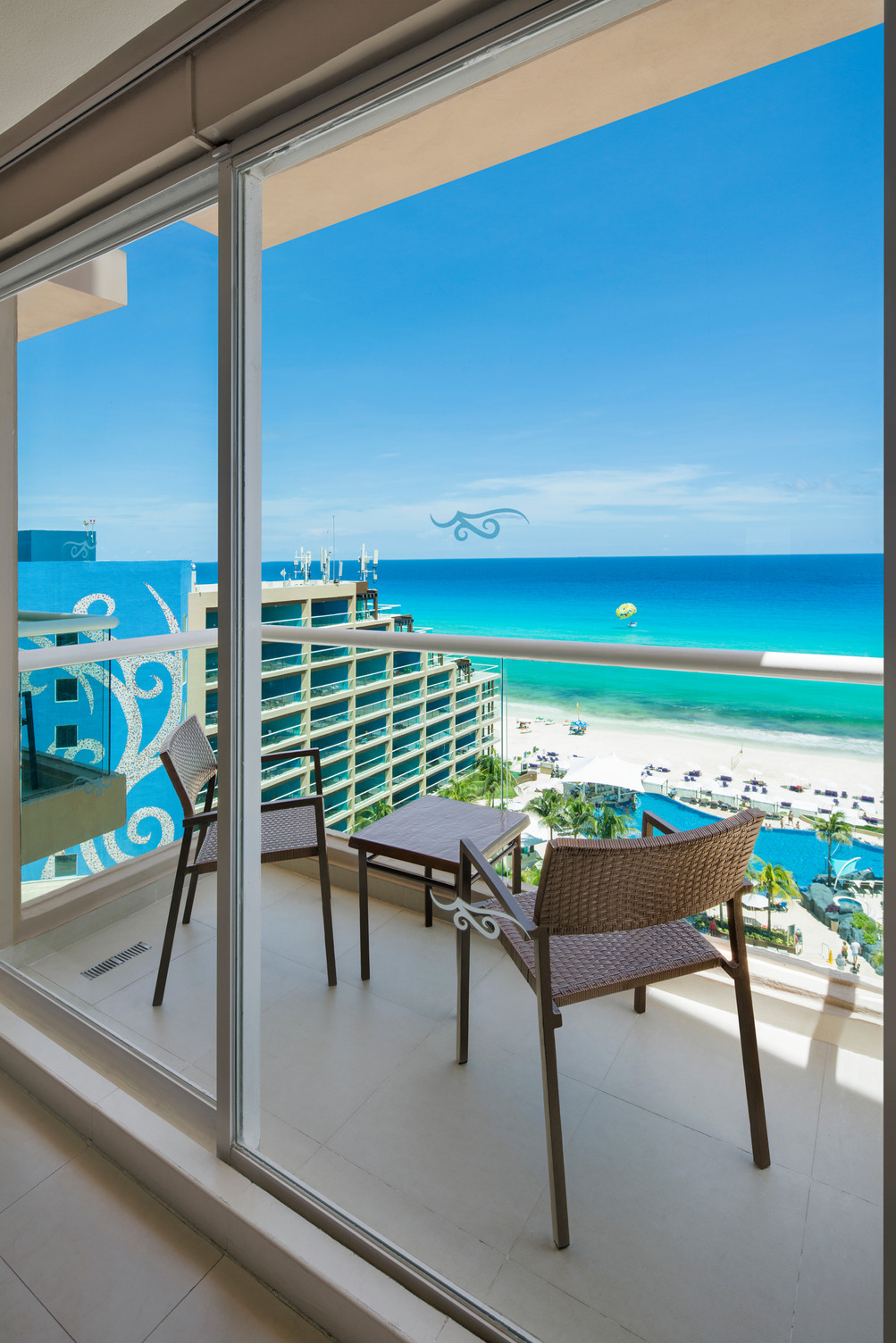 Sunny balcony with two chairs overlooking a vibrant blue sea and sandy beach, seen through a glass door.