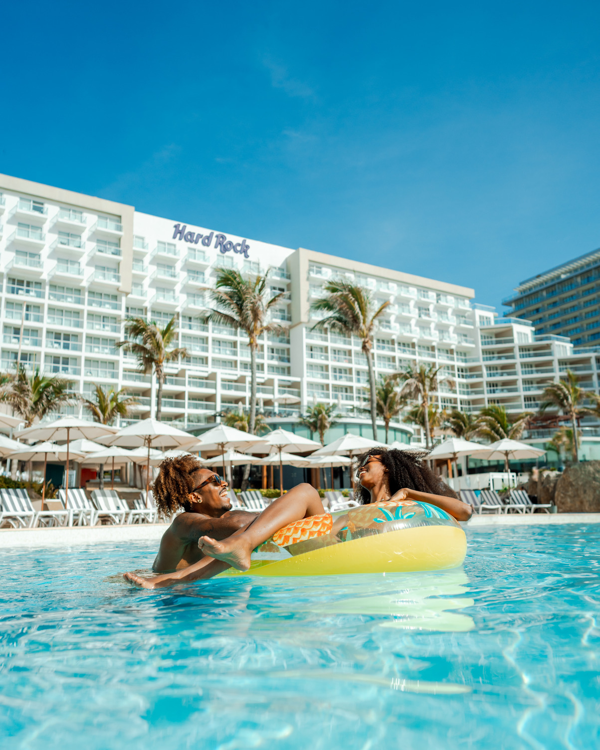 Couple relaxing on a float in a tropical pool at a resort with "Hard Rock" sign in the background.