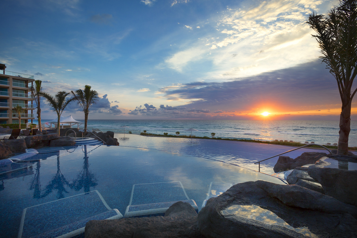 Infinity pool at sunset overlooking the ocean, flanked by palm trees and a hotel on the left, in Cancun.