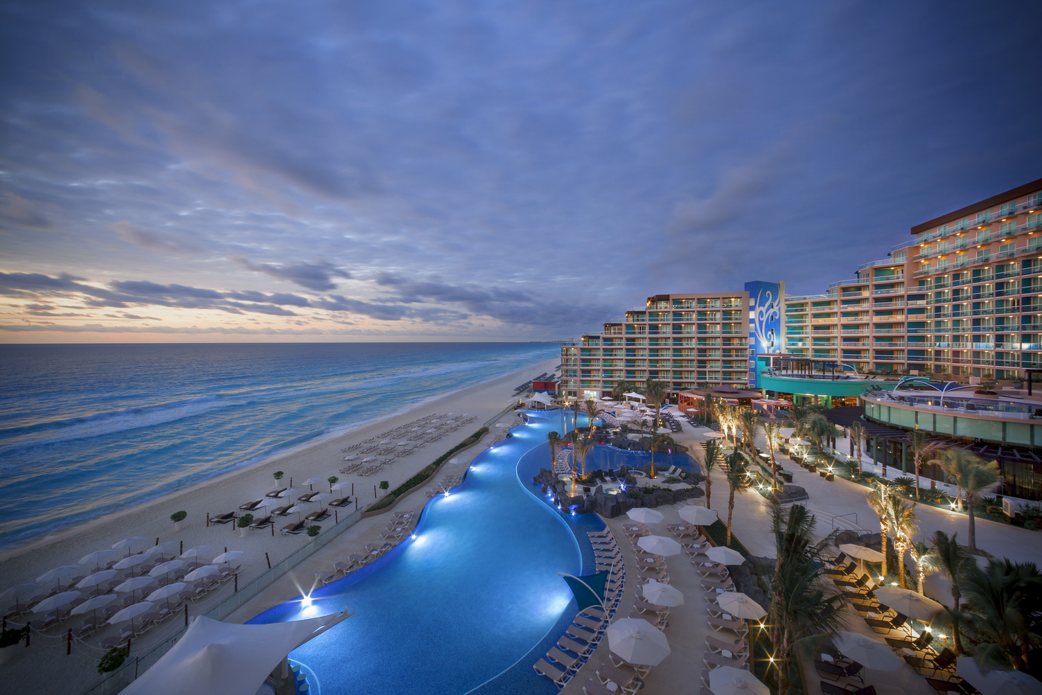 Vista al atardecer del área de alberca y playa del Hard Rock Cancun con el océano, camastros y edificios del resort al fondo.