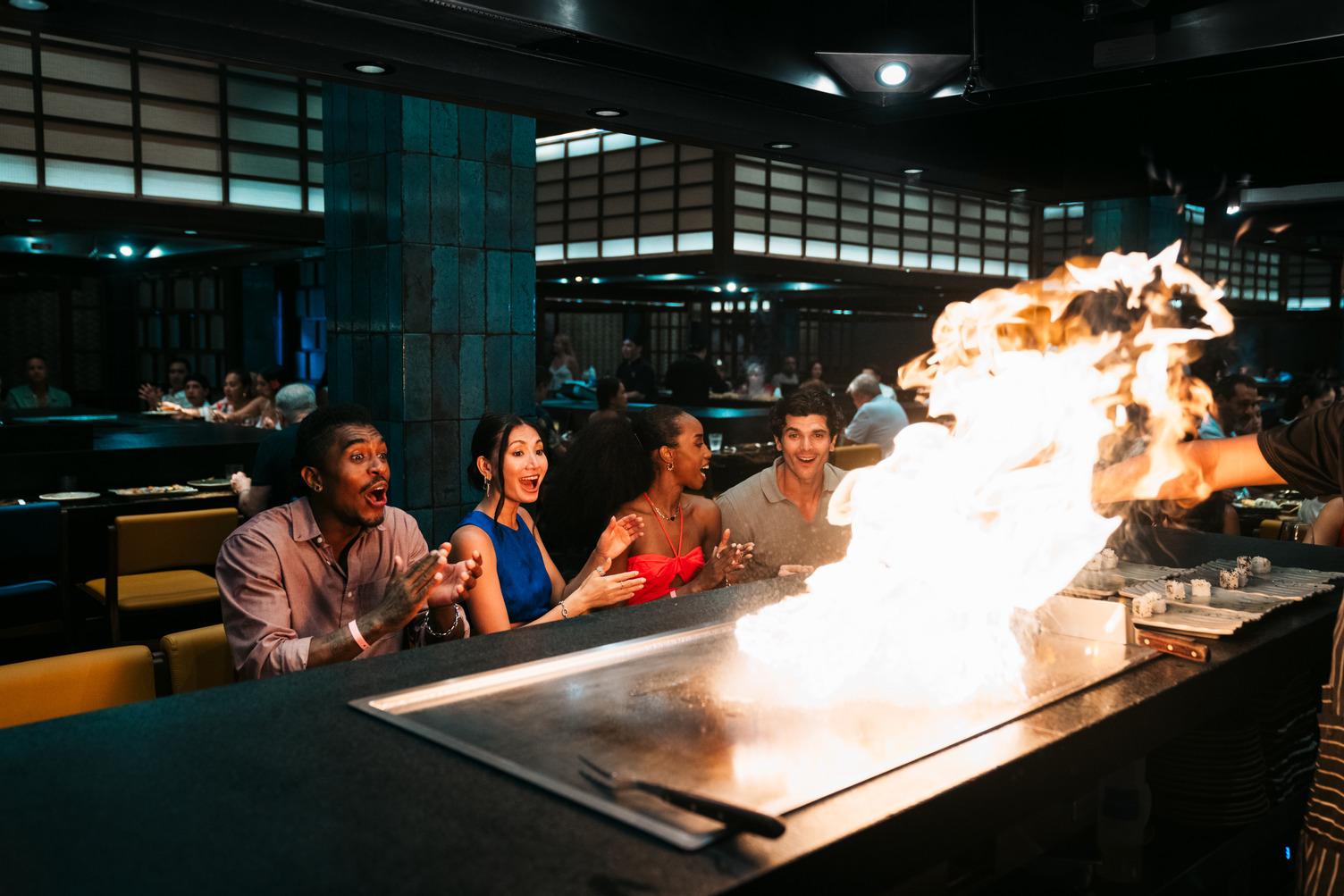 Grupo de cuatro personas mirando con emoción a un chef de hibachi crear una exhibición de fuego en la estación de parrilla de