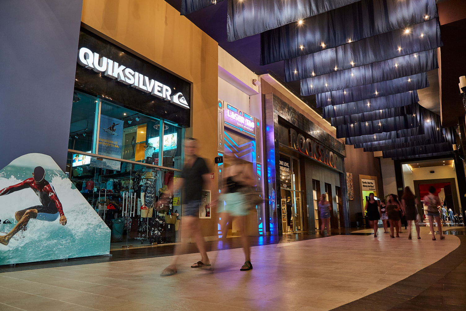 Personas caminan frente a las tiendas en el bulevar interior de Hard Rock Punta Cana, con techos iluminados y escaparates.