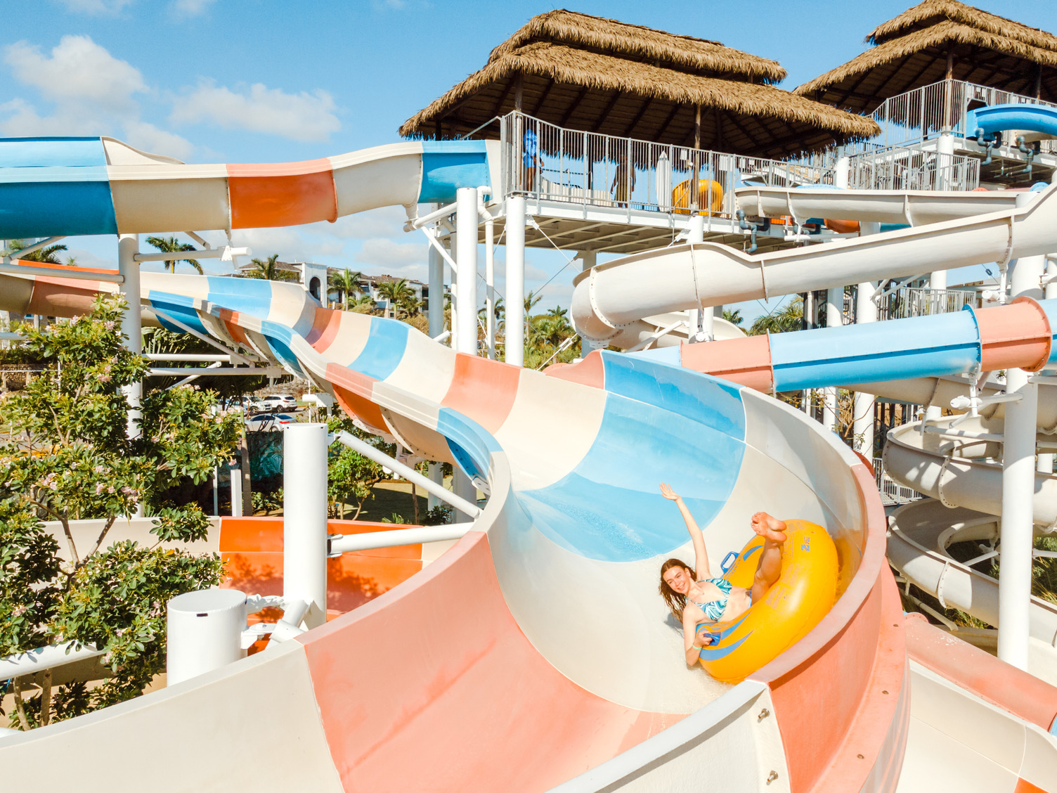 Water park with person in a yellow inflatable on a colorful slide, surrounded by trees and thatched-roof structures.