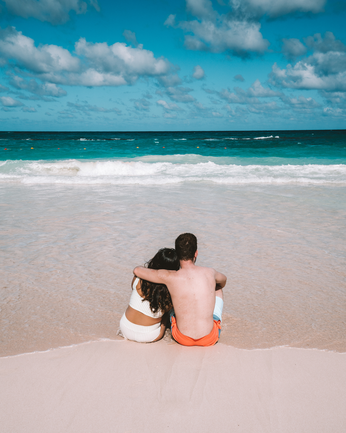 Couple sitting on sandy beach facing turquoise ocean, with blue sky and clouds in the background.