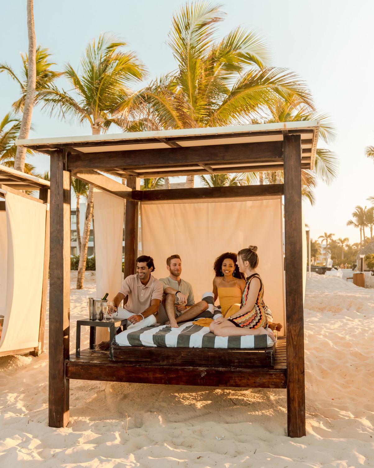 Cuatro personas sentadas en una cama de cabaña bajo un dosel de madera en una playa de arena con palmeras a la luz del día.