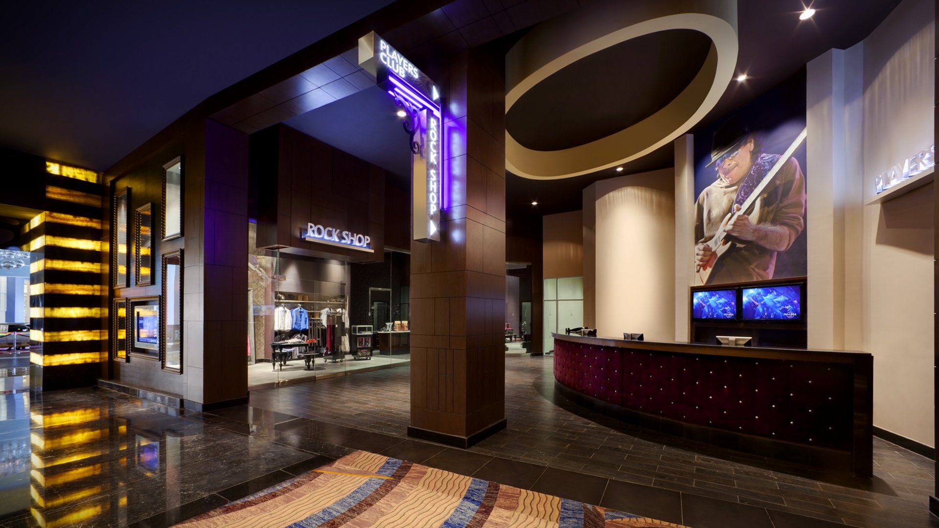 Área del lobby de un resort con la tienda Players Club Rock Shop, decoración moderna, letreros iluminados y un mural temático de guitarra.