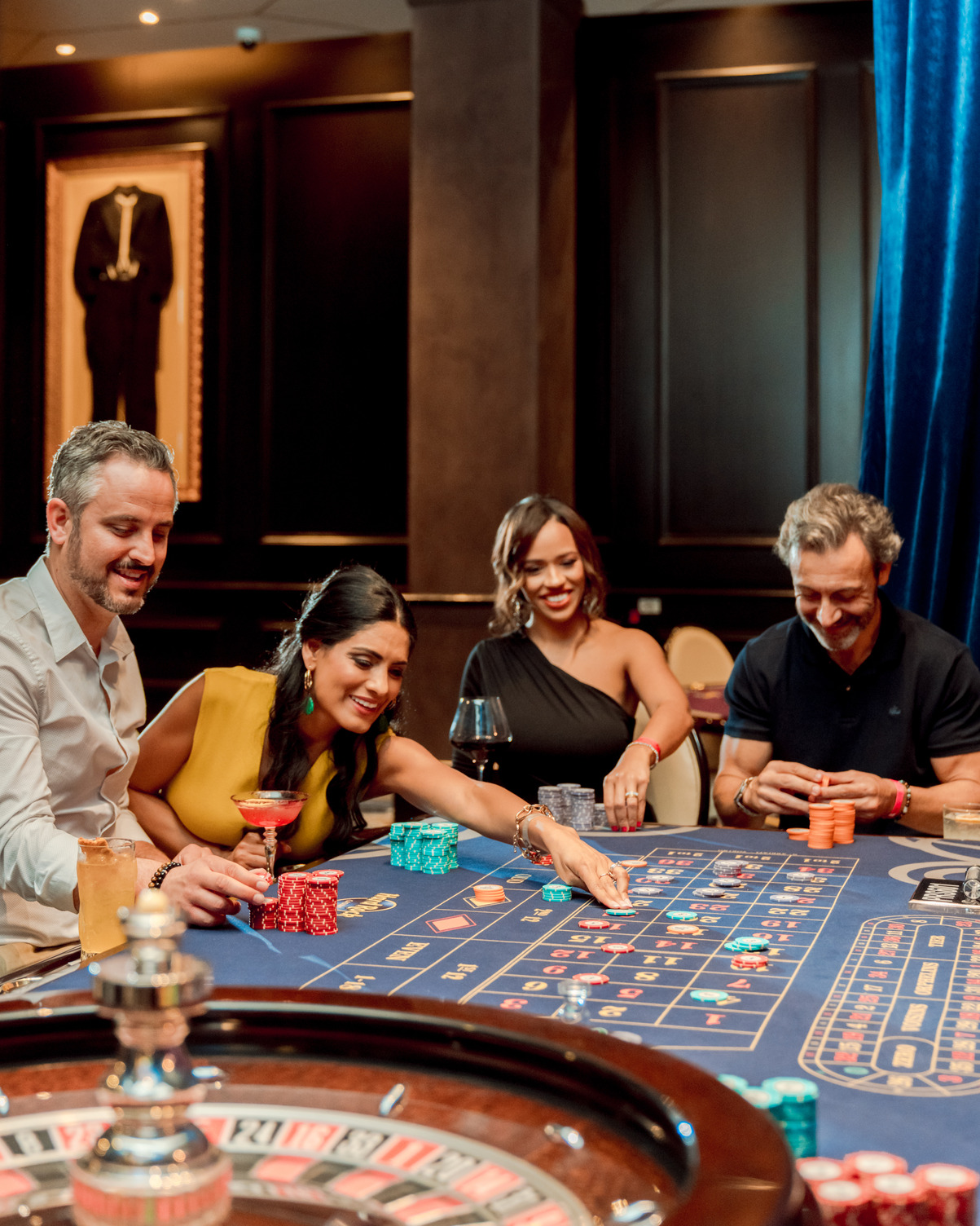 Personas disfrutando una partida en una mesa de ruleta del casino, con bebidas y fichas, en un ambiente animado.