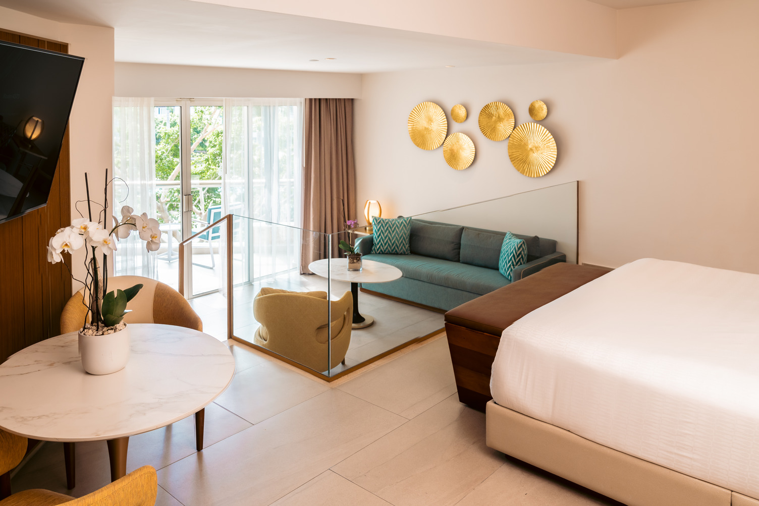 Elegant hotel room with a king bed, sofa bed, balcony view, wall art, and a small dining area with a potted orchid.
