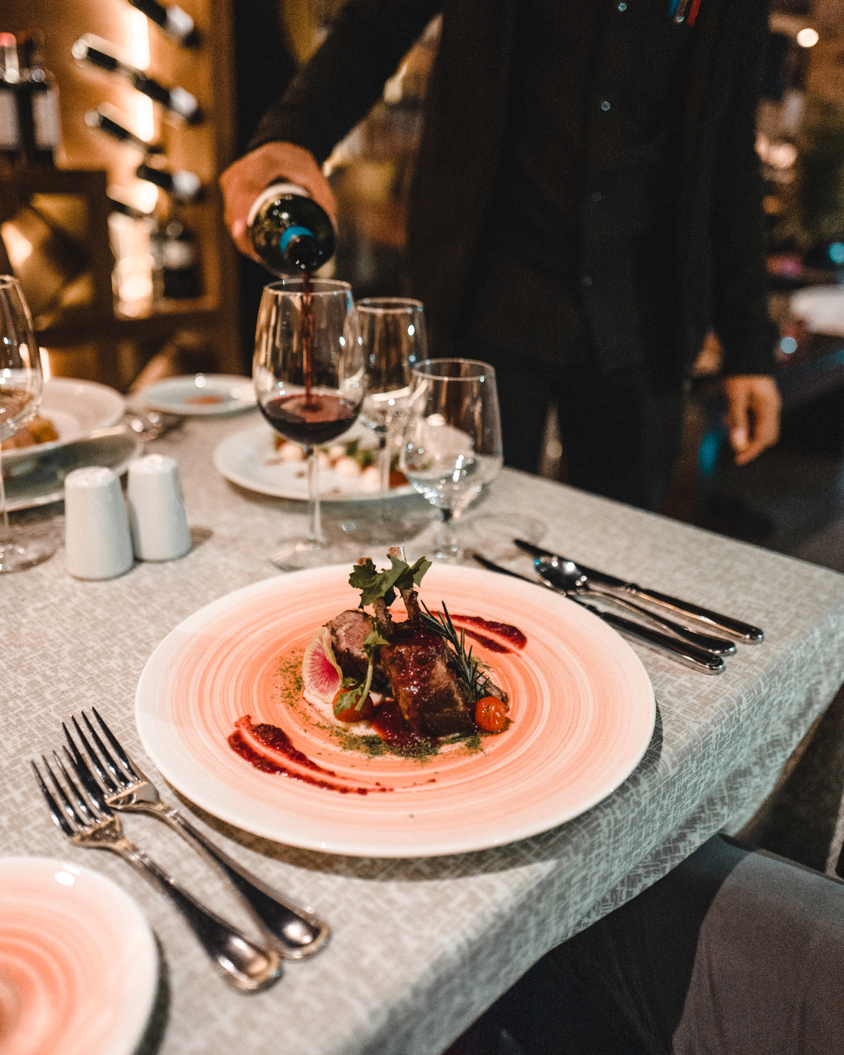 Vino siendo vertido en una copa junto a un platillo gourmet en la mesa de un restaurante con un ambiente moderno y elegante.
