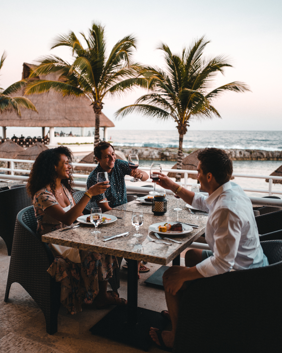 Tres personas cenando al aire libre en un restaurante junto al mar, brindando con copas de vino, con palmeras al fondo.