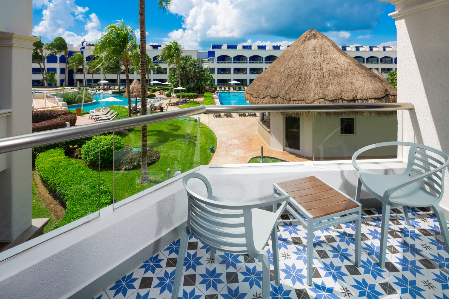 Balcón con azulejos de patrones azules, dos sillas, mesa, con vista a la alberca, palmeras y edificios del resort bajo un cie