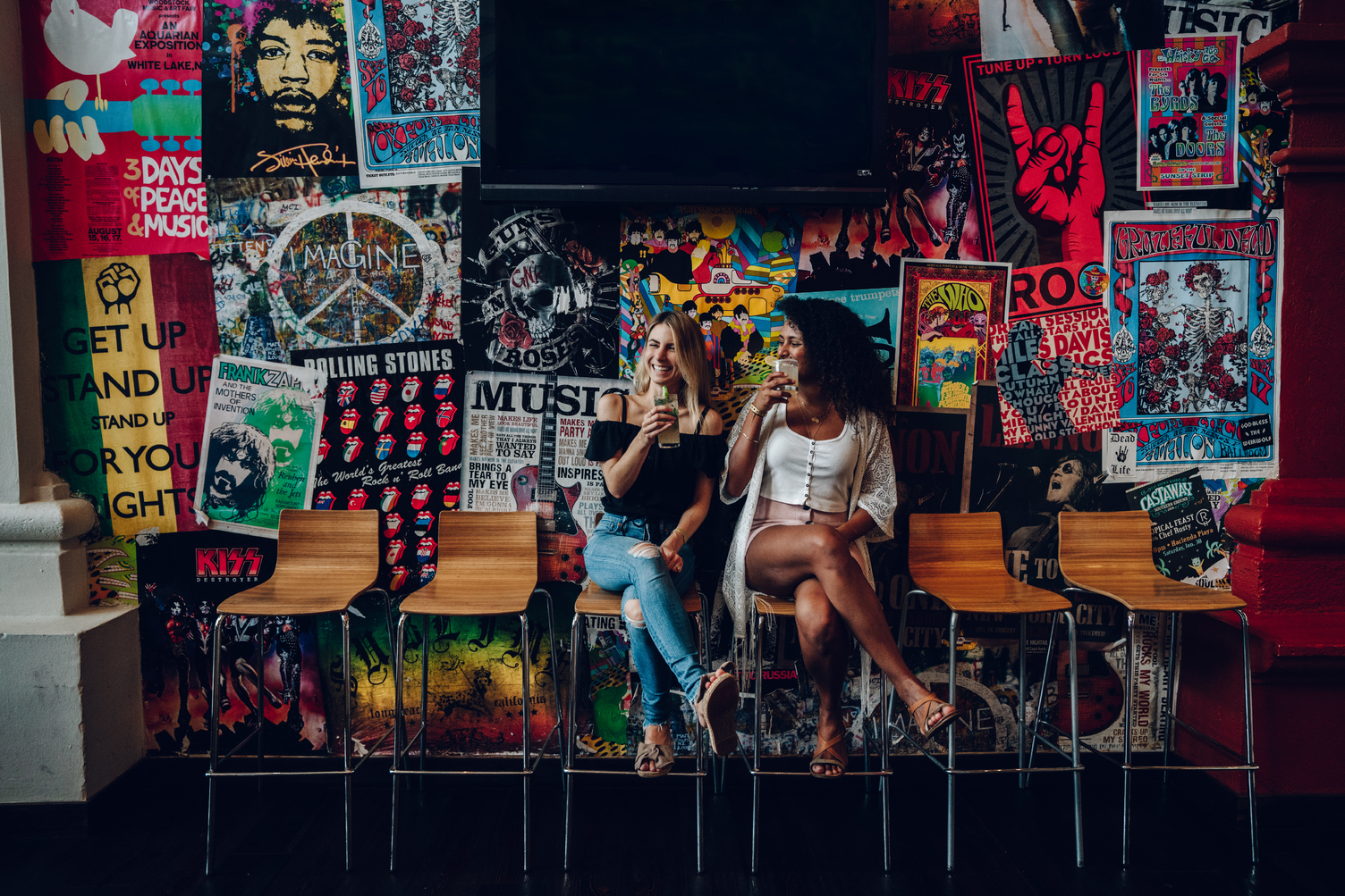 Dos mujeres disfrutando de bebidas en un bar con una pared de pósters coloridos de música rock detrás de ellas.