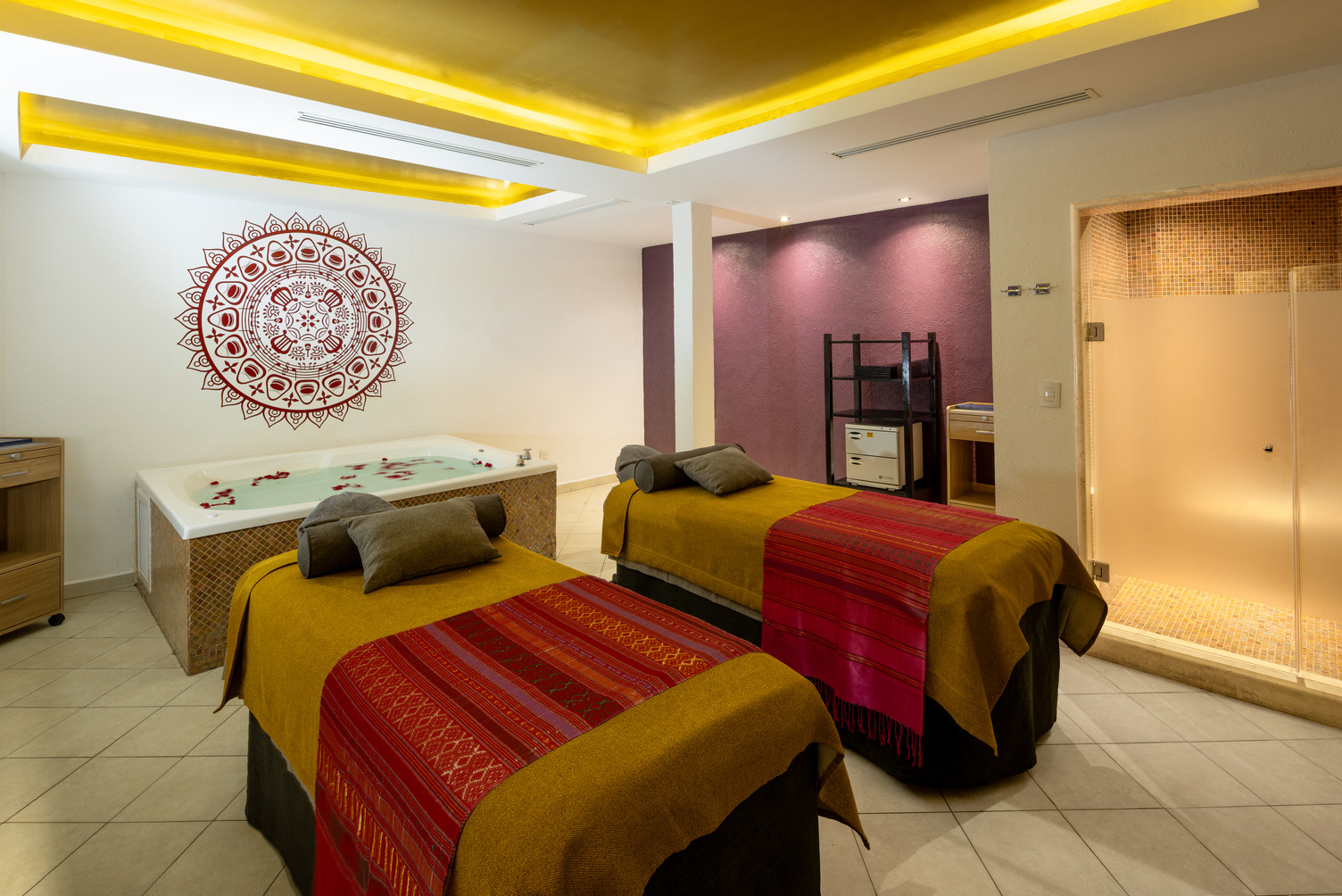 Spa room with two massage tables, colorful blankets, a hot tub with flower petals, and serene lighting.