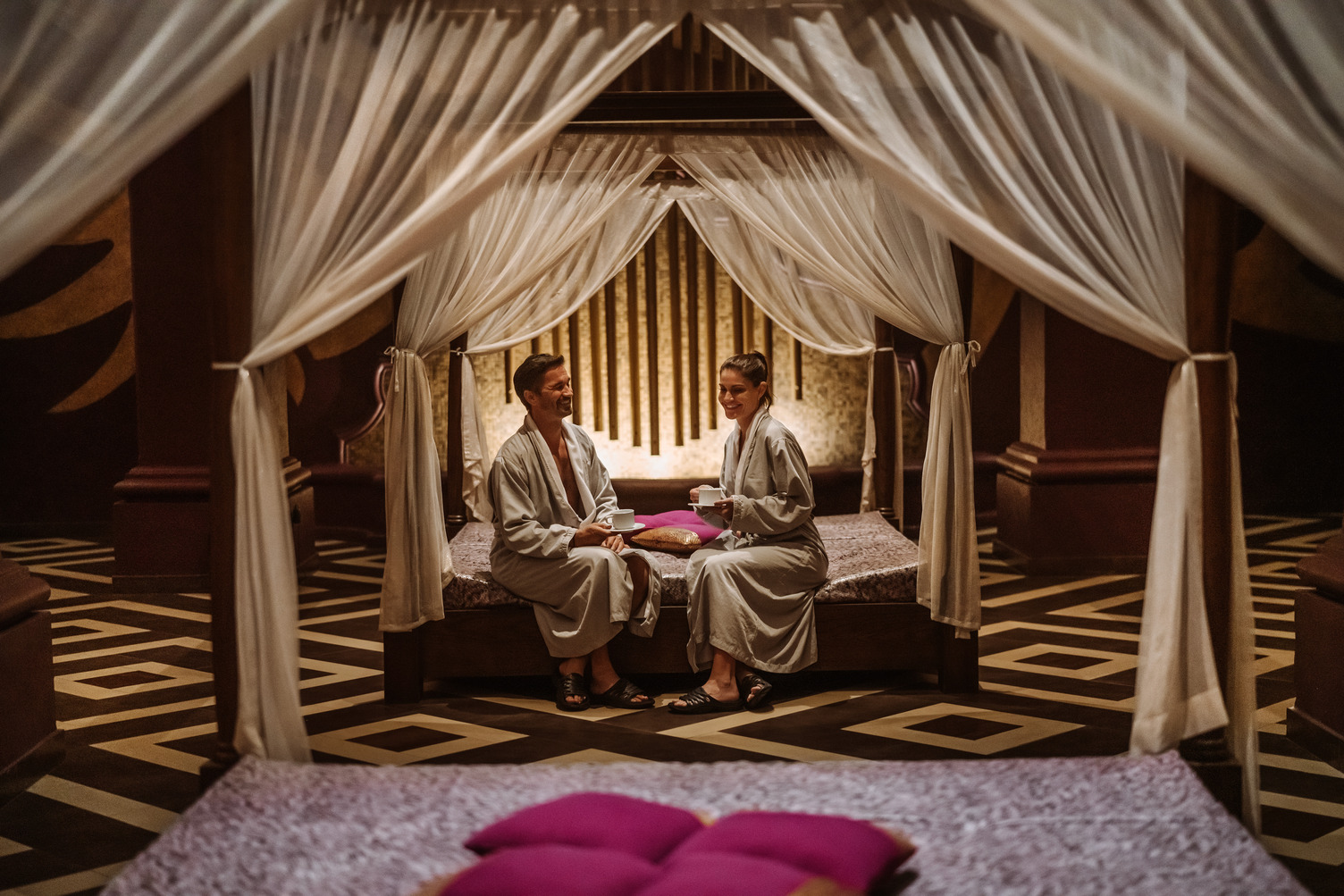 A couple in robes relaxes on a bed with draped canopy, each holding a cup, in a serene spa setting.