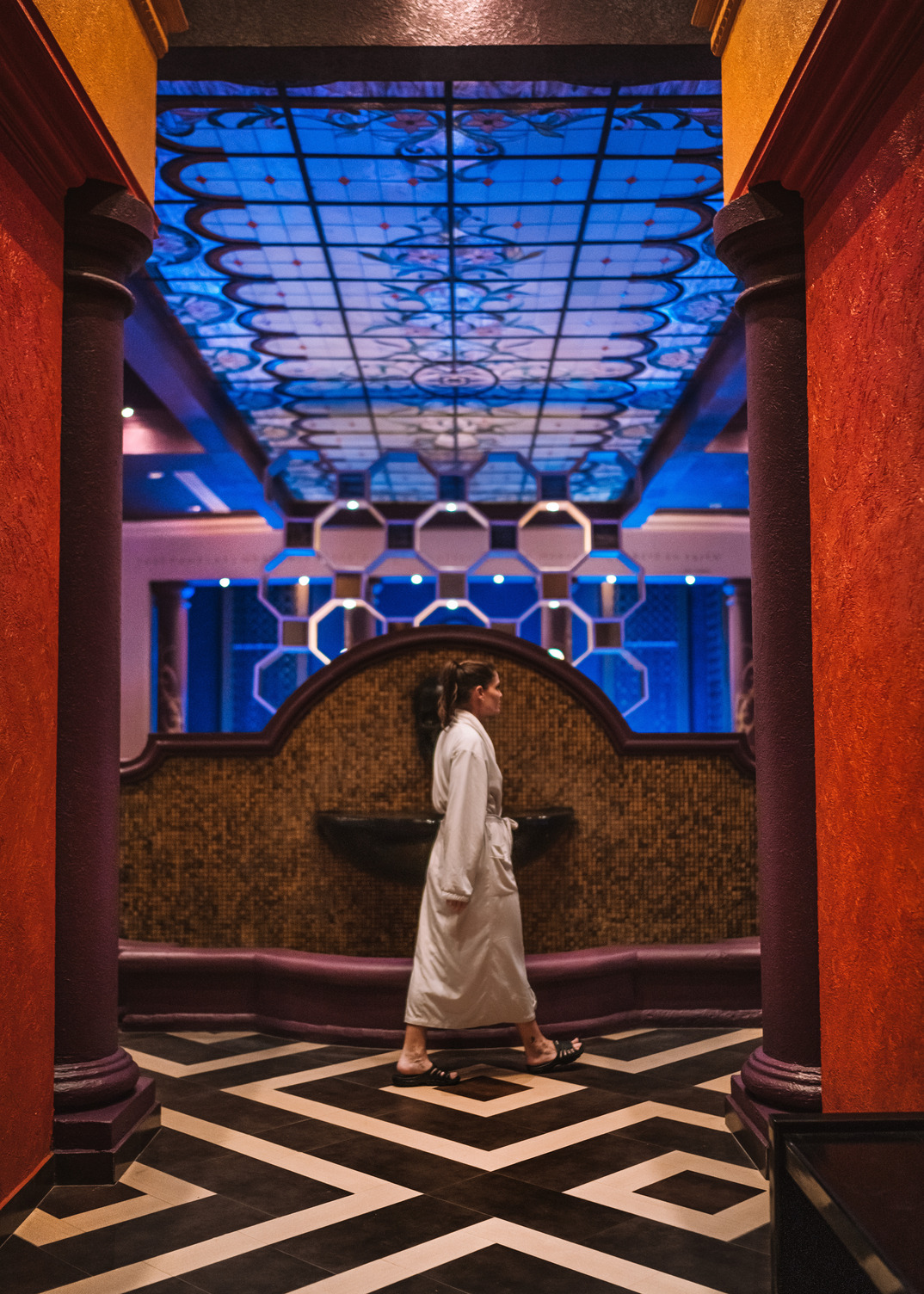 Person in a robe walks through ornate spa hallway with colorful ceiling and geometric floor pattern.
