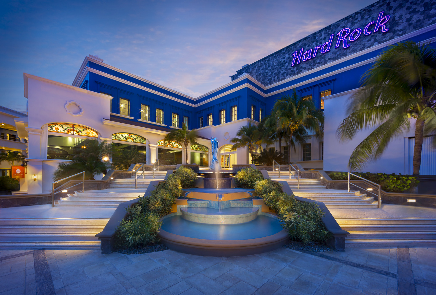 Escalera al anochecer que conduce a la entrada de Hard Rock Riviera Maya, enmarcada por palmeras e iluminación vibrante.