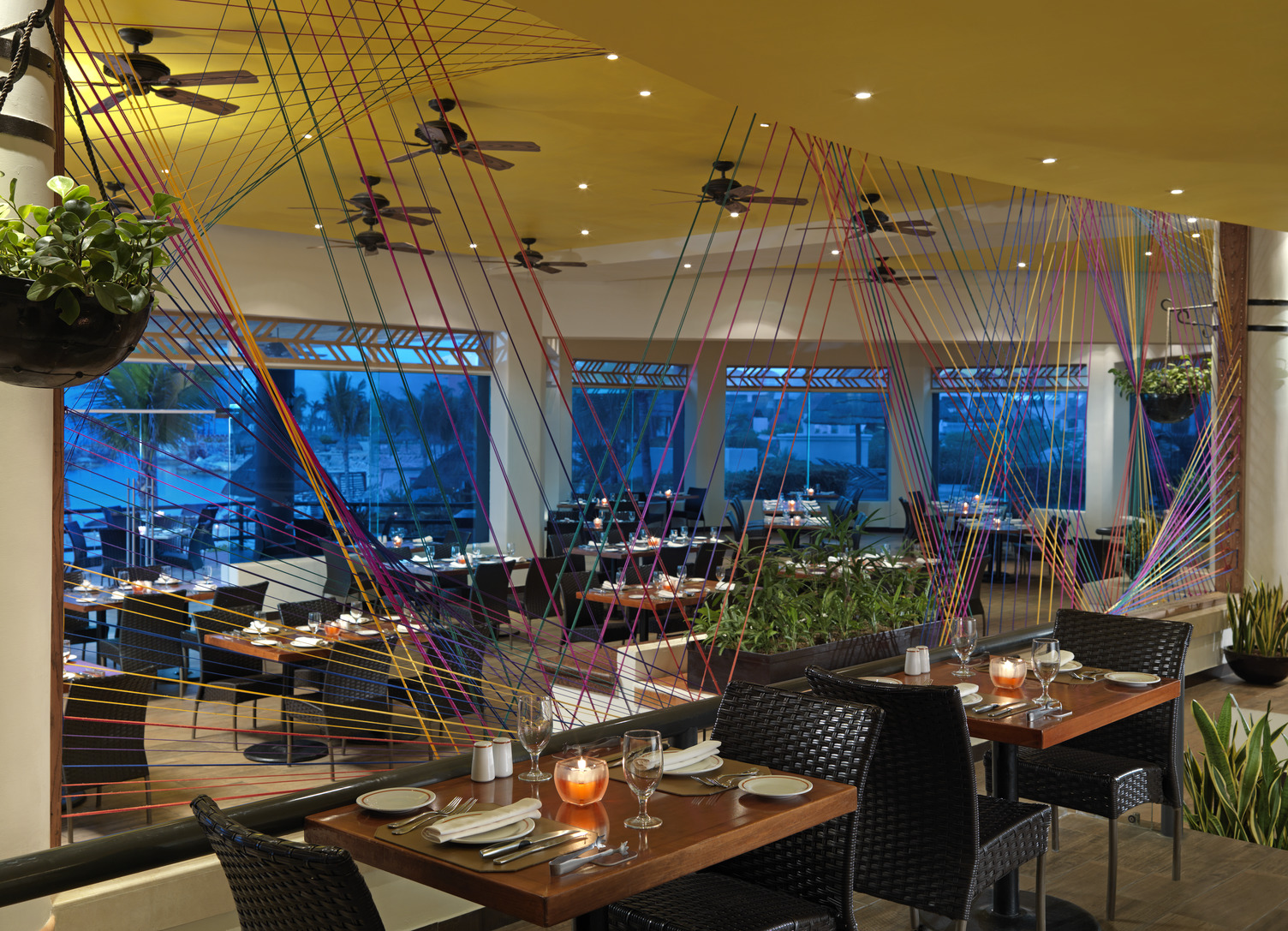 Elegante ambientación de restaurante con arte de hilos vibrantes, mesas iluminadas con velas, sillas de mimbre y grandes vent